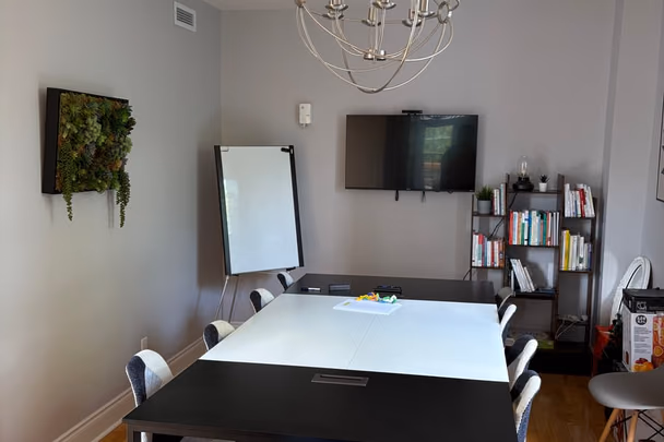 Une salle de conférence avec une longue table blanche, des chaises, un tableau blanc, un écran mural, une bibliothèque et un cadre de mousse végétale.