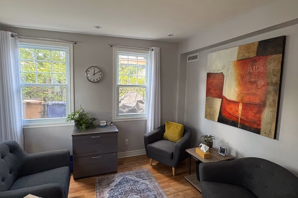 Un bureau avec deux fauteuils gris de part et d'autre d'un guéridon, une grande toile abstraite rouge et beige, un tapis ancien, un classeur de bois et deux fenêtres donnant sur les arbres.