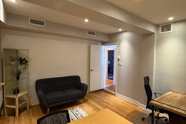 Une salle de travail avec un canapé vert foncé contre un mur de brique peint en blanc, une grande table de bois clair, une chaise de bureau et une vitrine garnie de plantes.