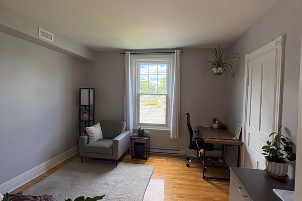 Un bureau clair avec un petit canapé gris sous la fenêtre, une table de travail avec sa chaise, un tapis épais, une plante suspendue et un plancher de bois blond.