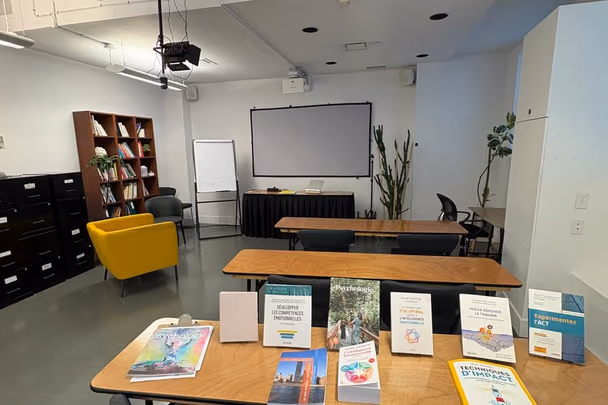 La salle de groupe avec deux longues tables de bois, des chaises, un écran de projection, une bibliothèque et un fauteuil jaune.