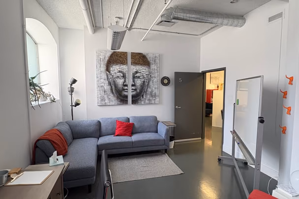 Un bureau avec un canapé d'angle gris, une grande toile représentant un visage de Bouddha, un fauteuil rouge et un tableau blanc sur roulettes.