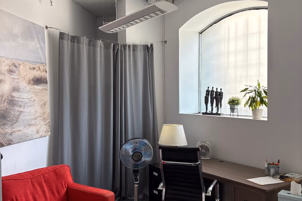 Un bureau avec un fauteuil rouge devant un rideau gris, une table de travail en bois, une chaise de bureau et un ventilateur sur pied.