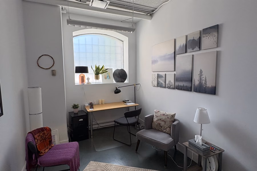 Un bureau clair avec une table de travail contre le mur, un fauteuil gris, un tapis à motifs concentriques et un triptyque photographique de paysages.