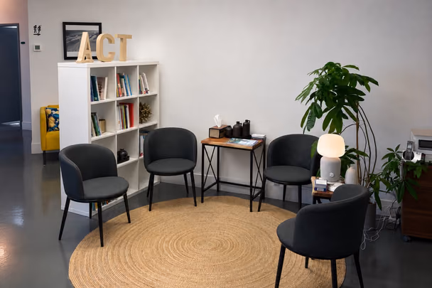 La salle d'attente avec quatre fauteuils gris autour d'une petite table d'appoint, un tapis rond de jute, une bibliothèque blanche à gauche, une grande plante et une lampe à droite, devant un mur blanc nu.