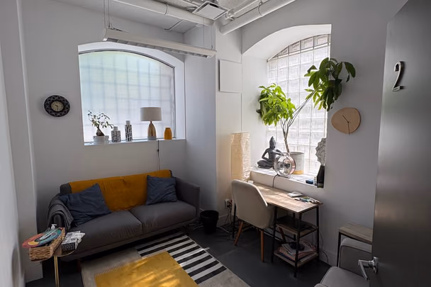 Un bureau avec un canapé gris, un tapis jaune posé sur un tapis rayé, une table de travail claire et une plante haute devant la fenêtre.