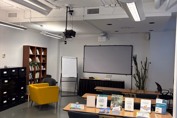 La salle de formation avec deux longues tables de bois, un écran de projection, un tableau de conférence, une bibliothèque et un fauteuil jaune.