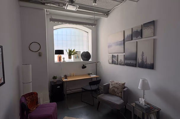Un bureau avec une table de travail sous une fenêtre cintrée, une chaise, une série de photographies de paysages au mur, un fauteuil gris et un fauteuil violet.