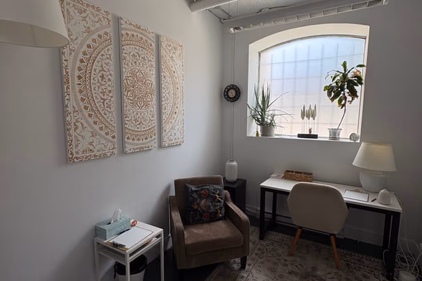 Un bureau avec un fauteuil de cuir brun, une table de travail claire sous une fenêtre cintrée, un triptyque de panneaux sculptés au mur et des plantes sur l'appui.