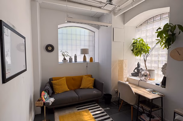 Un bureau vu depuis la porte avec un canapé gris à coussins jaunes, un tapis rayé noir et blanc posé sur un tapis jaune, une table de travail sous les fenêtres cintrées et une statuette assise sur l'appui.